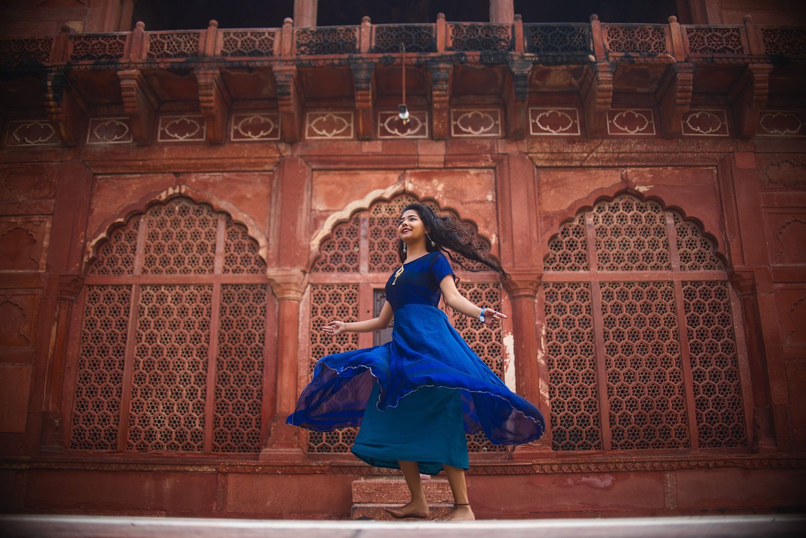 A woman in a blue dress dances joyfully in front of a historic monument in Agra, India.